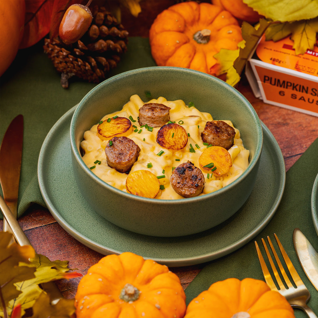 Bowl of macaroni and cheese with sausages and butternut squash on a festive table with pumpkins and autumn decorations.