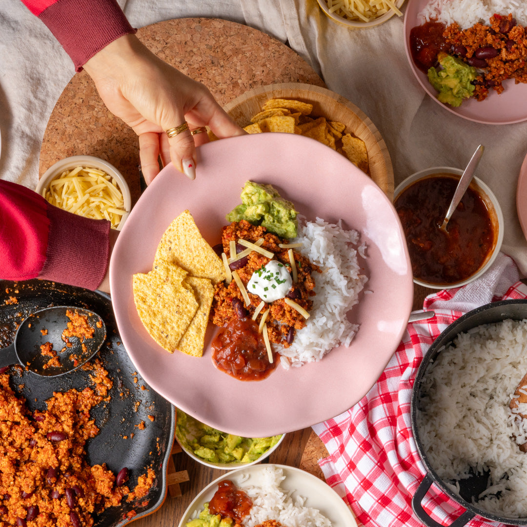 A pink plate filled with a Mexican-style meal, including white rice, red salsa, guacamole, shredded cheese, and tortilla chips, held by a person's hand in a cozy kitchen setting with other food bowls and plates around it.